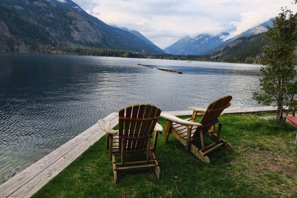 Two chairs at the waters edge with mountains surrounding the water.