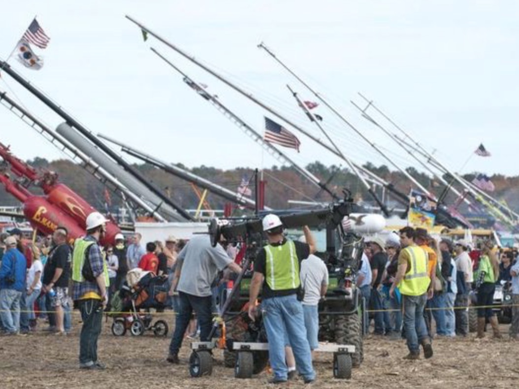 Line of punkin chunkin machines in a line with crowds gathered. 