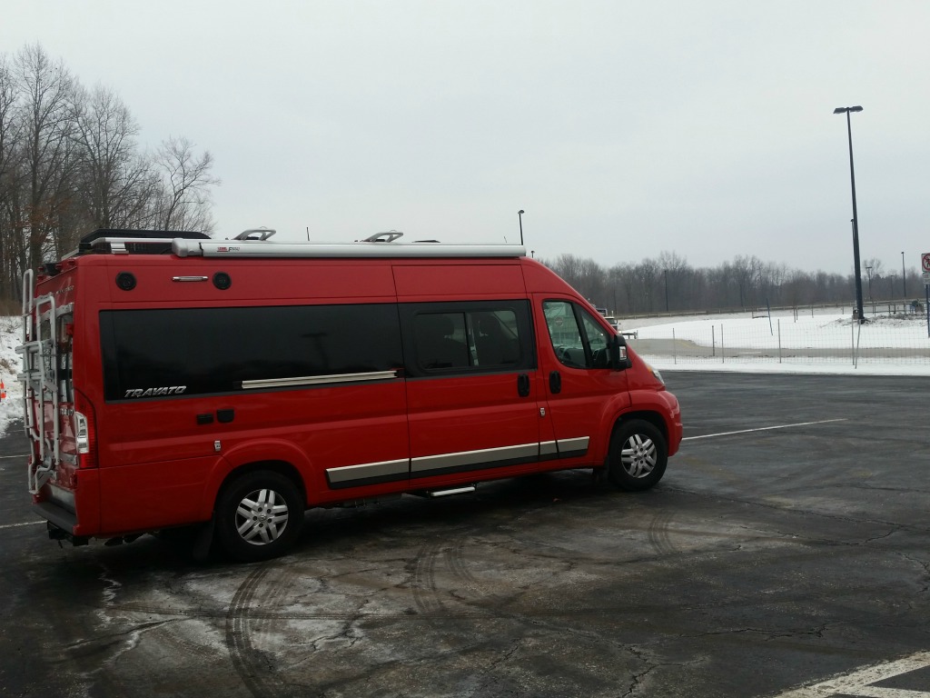 Red Winnebago Travato in parking lot with snow covering the surrounding ground.