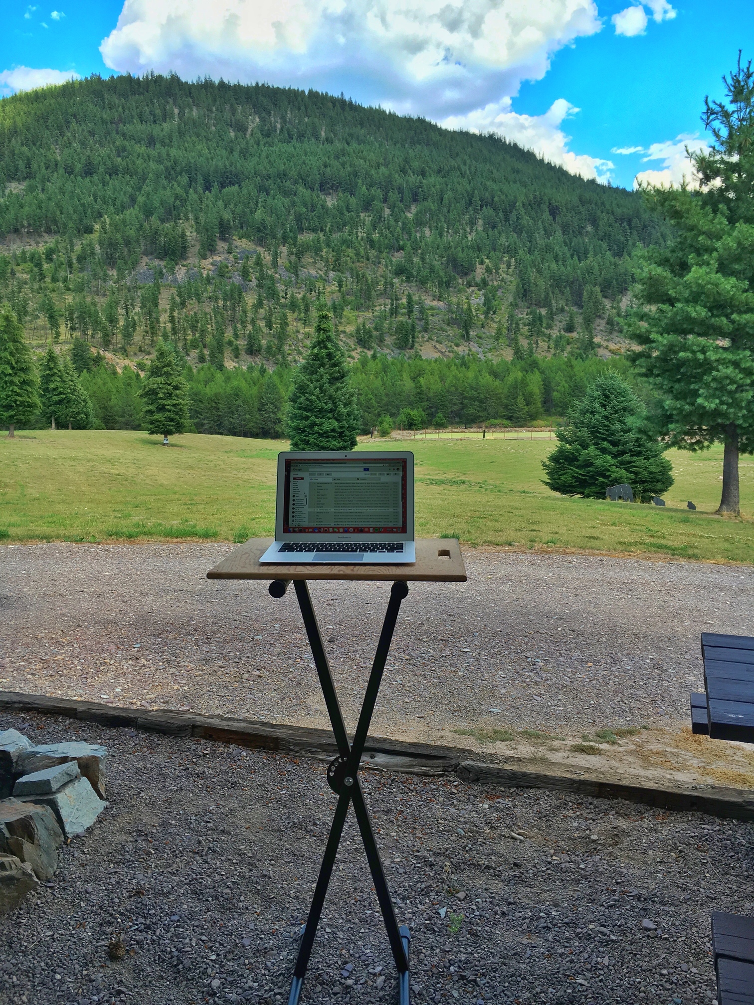 Computer at makeshift standing desk outside motorhome with beautiful tree filled hillside ahead.