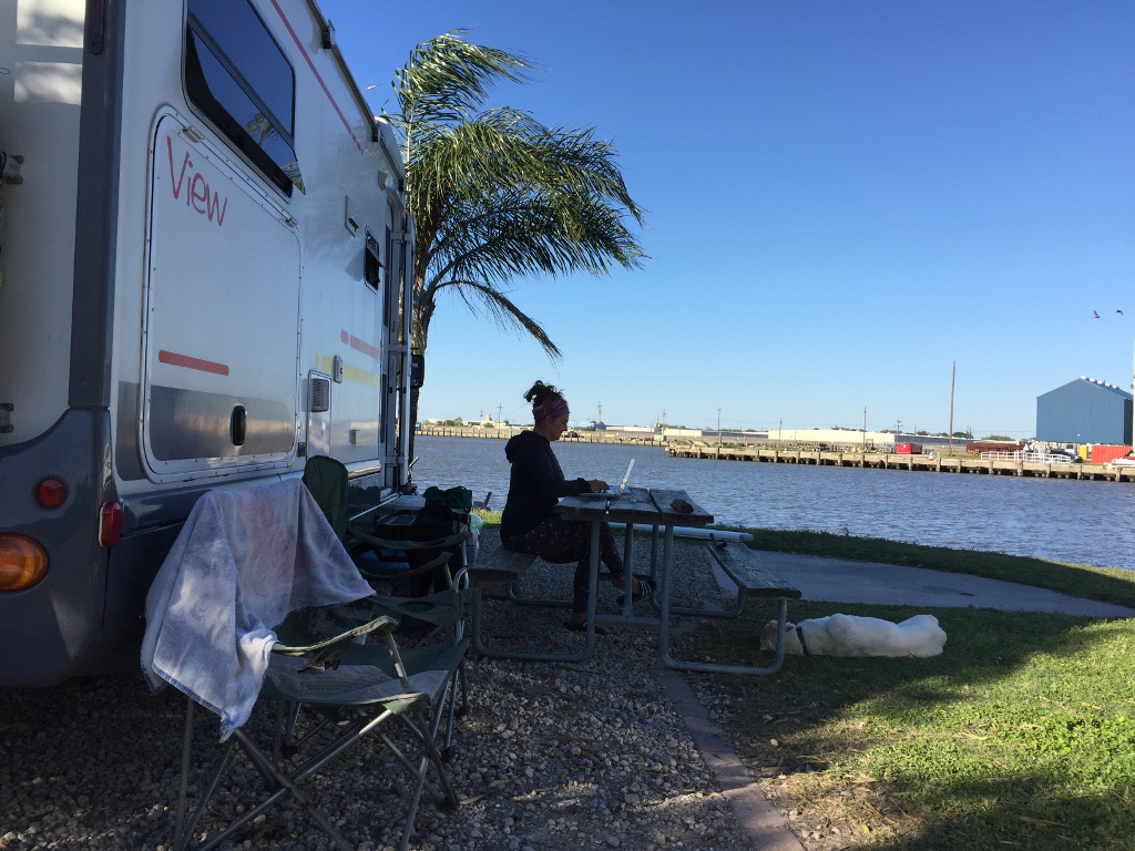 Woman working on laptop at picnic table outside RV next to the water.