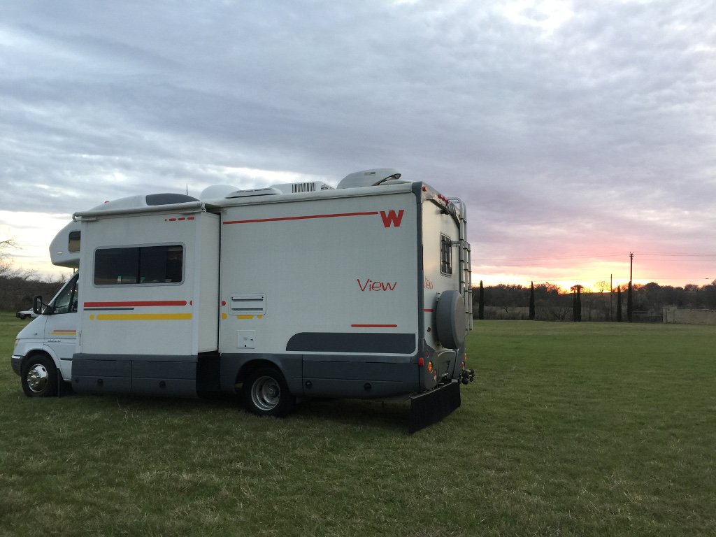 Winnebago View parked in grassy field with sun setting over the horizon.