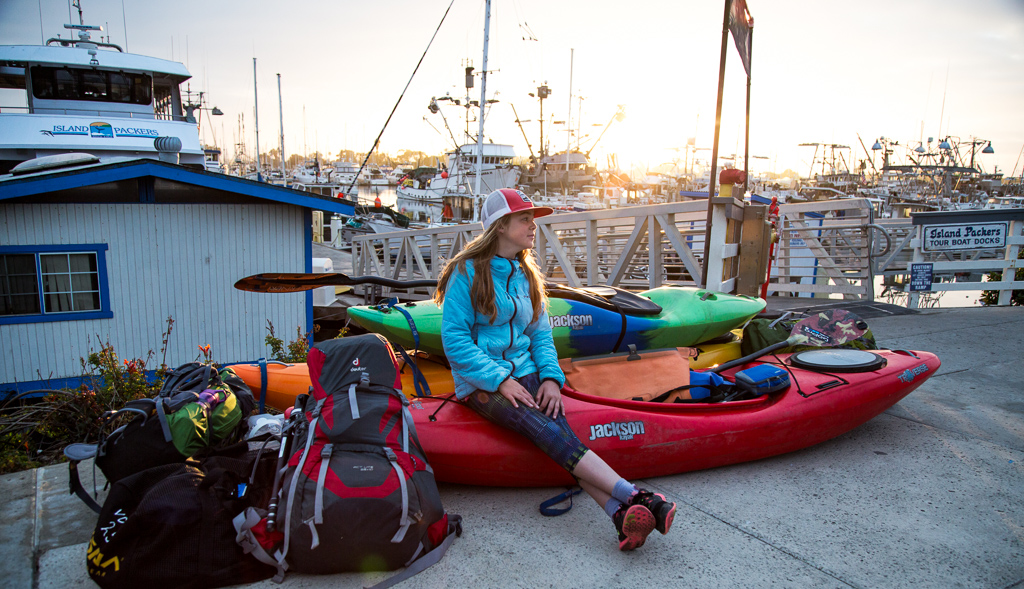 Abby sitting among the family's kayaks and backpacks with boats docked in the harbor behind her.