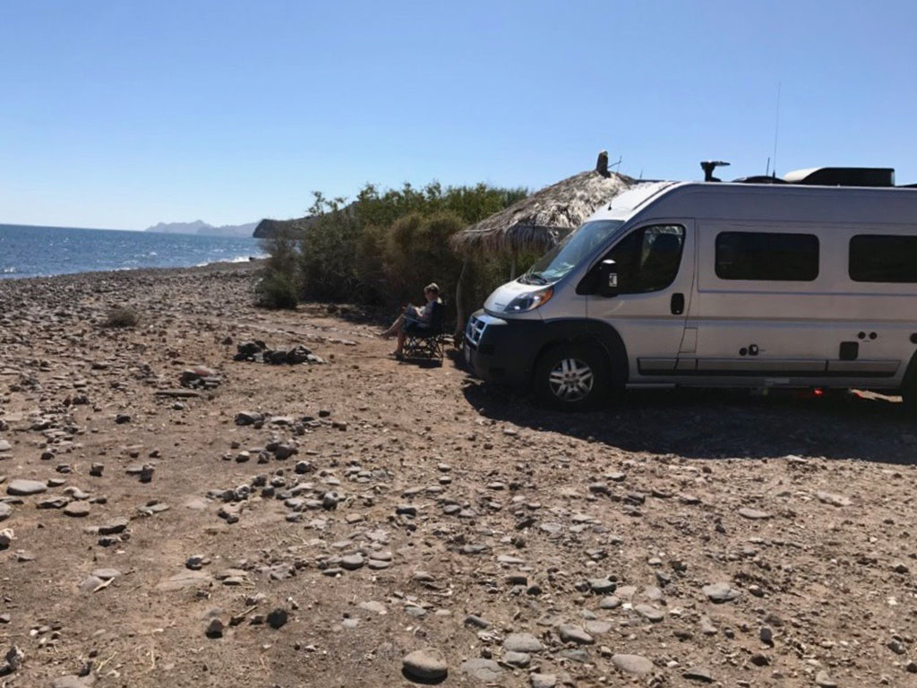 Woman sitting on the beach in front of her Winnebago Travato reading a book.