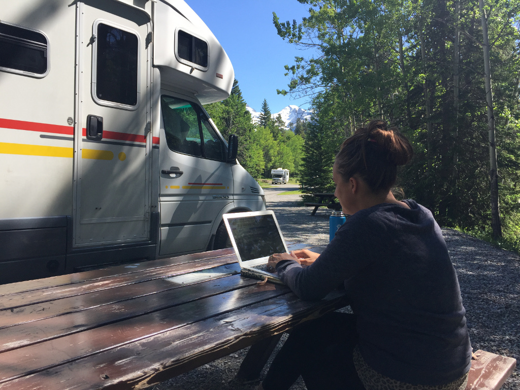 Woman on a computer at a picnic table outside her Winnebago View parked at campsite nestled in the trees and mountains peeking above the trees in the distance.