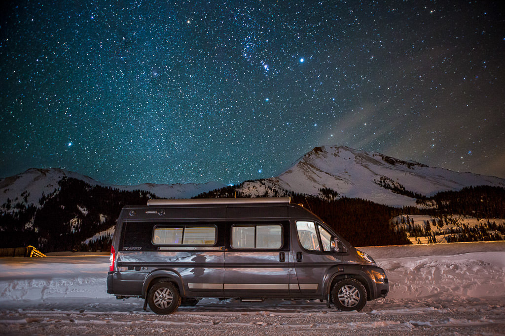 Winnebago Travato parked on snow covered ground with mountains in the background under a star filled night sky.